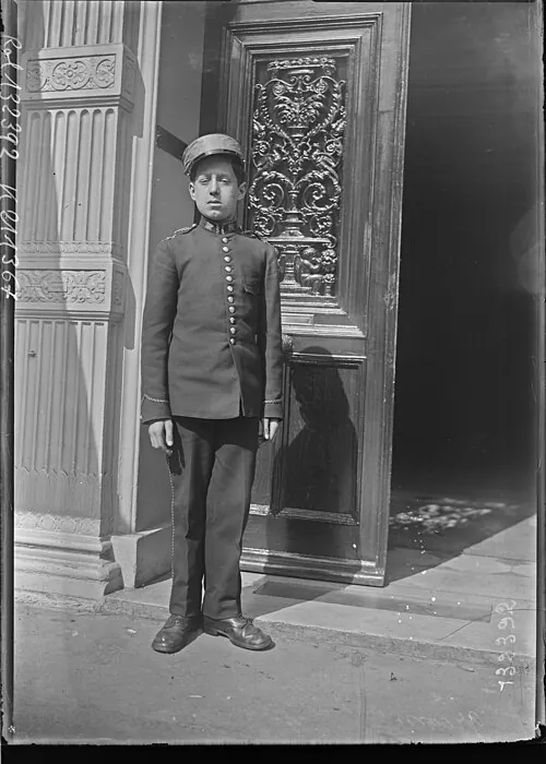 A young hotel groom in a short, fitted, buttoned uniform jacket and pillbox cap, standing by a carved door